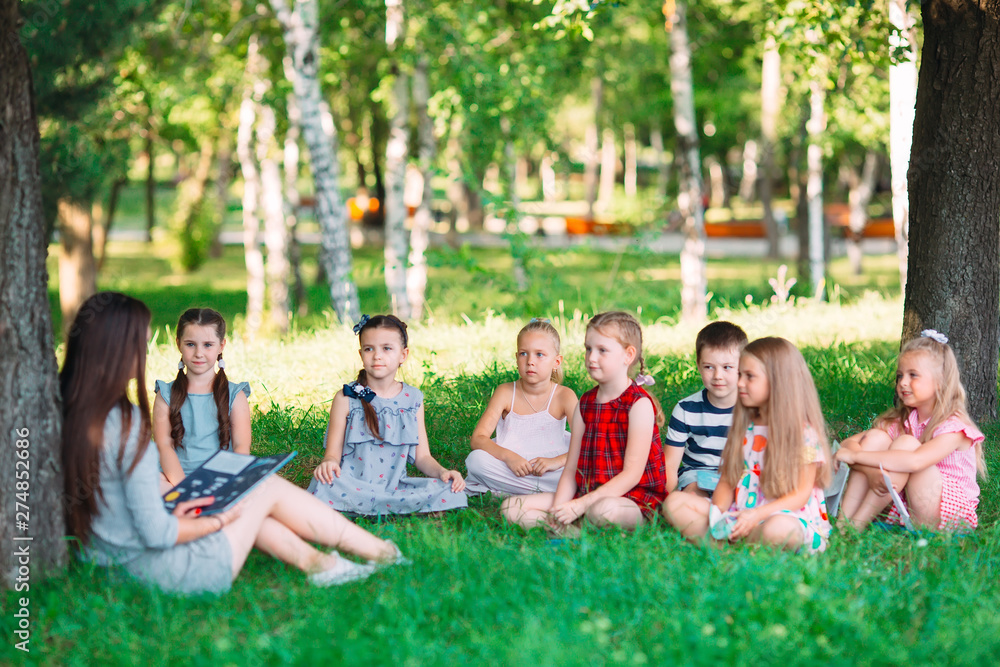 Children and education, young woman at work as educator reading book to boys and girls in park. Children and education, young woman at work as educator reading book to boys and girls in park.