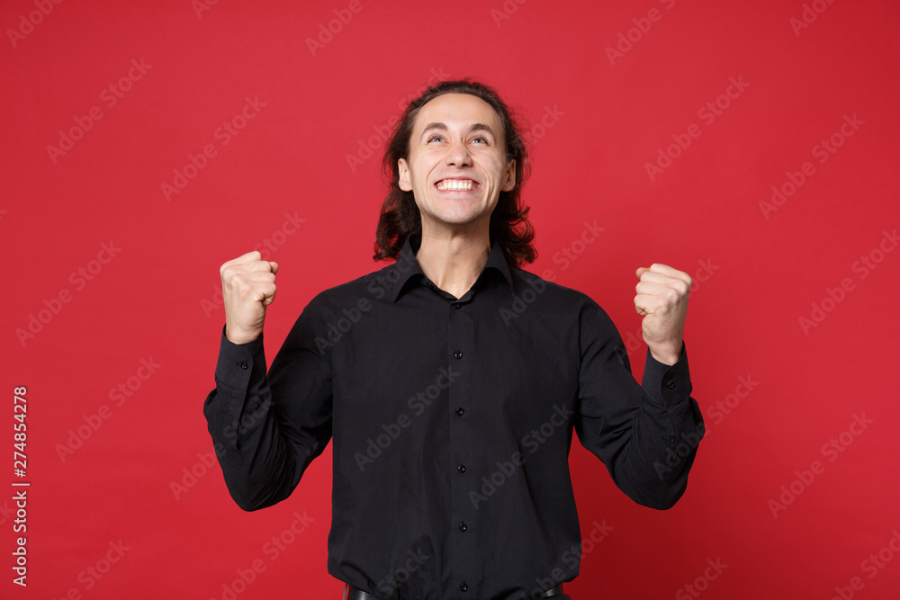 Handsome stylish young curly long haired man in black shirt posing isolated on red wall background studio portrait. People sincere emotions lifestyle concept. Mock up copy space. Doing winner gesture. Handsome stylish young curly long haired man in black shirt posing isolated on red wall background studio portrait. People sincere emotions lifestyle concept. Mock up copy space. Doing winner gesture.