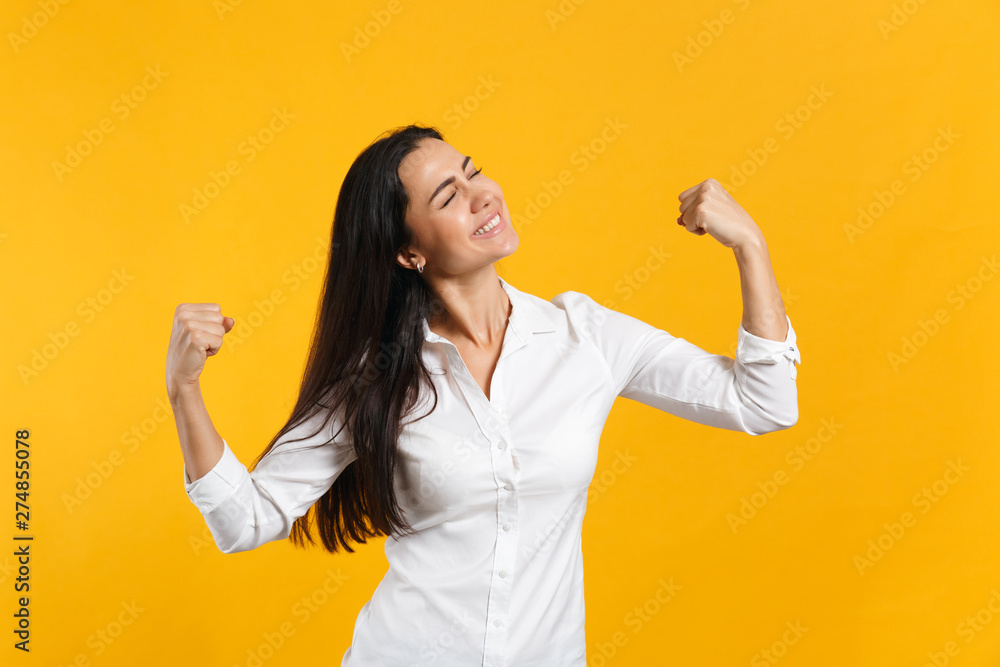 Portrait of stunning smiling young woman in white shirt keeping eyes closed, clenching fists like winner isolated on yellow orange background in studio. People lifestyle concept. Mock up copy space. Portrait of stunning smiling young woman in white shirt keeping eyes closed, clenching fists like winner isolated on yellow orange background in studio. People lifestyle concept. Mock up copy space.