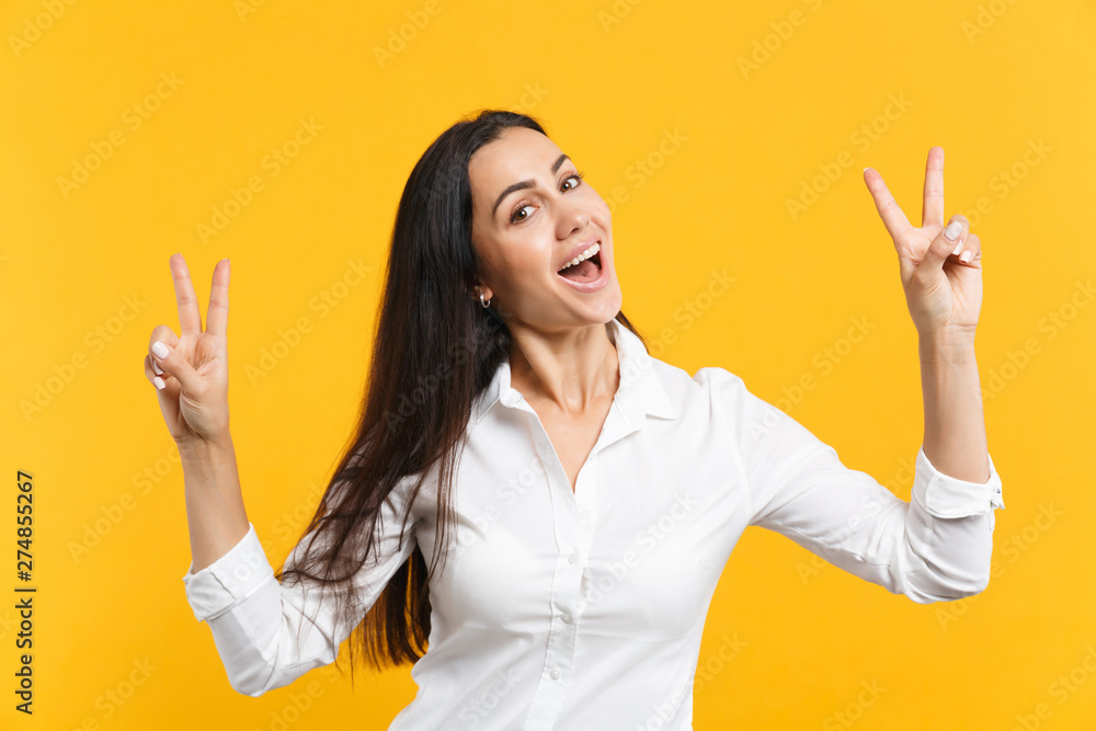 Portrait of cheerful funny young woman in white casual shirt looking camera, showing victory sign isolated on yellow orange wall background in studio. People lifestyle concept. Mock up copy space. Portrait of cheerful funny young woman in white casual shirt looking camera, showing victory sign isolated on yellow orange wall background in studio. People lifestyle concept. Mock up copy space.