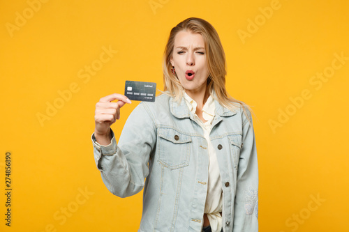 Portrait of surprised amazed young woman in denim casual clothes holding credit bank card isolated on bright yellow orange wall background in studio. People lifestyle concept. Mock up copy space.
