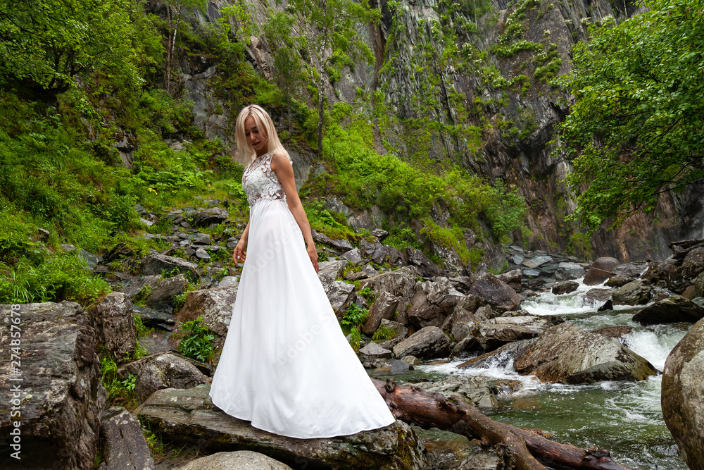 A young blonde girl standing in a half-turn and looking down at the hem of a boudoir dress in the mountains against a waterfall and stones like a ballerina on a warm summer day in the Altai green tree
