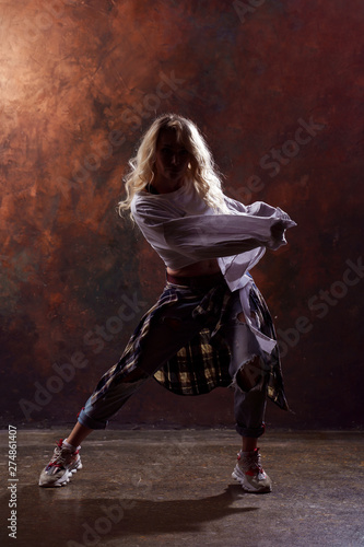 Photo of young dancing blonde girl in ripped jeans and sneakers on dark brown background in studio