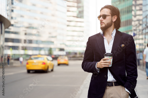 Photo of brunette with beard and earring in ear looking to side with glass in hand in city against background of tall buildings, cars