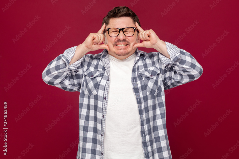 Headache pain or confusion. Portrait of confused middle aged business man in casual checkered shirt and eyeglasses standing and holding his painful head. studio shot, isolated on dark red background. Headache pain or confusion. Portrait of confused middle aged business man in casual checkered shirt and eyeglasses standing and holding his painful head. studio shot, isolated on dark red background.