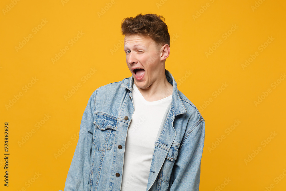 Portrait of cheerful young man in denim casual clothes blinking and looking camera isolated on yellow orange wall background in studio. People sincere emotions, lifestyle concept. Mock up copy space. Portrait of cheerful young man in denim casual clothes blinking and looking camera isolated on yellow orange wall background in studio. People sincere emotions, lifestyle concept. Mock up copy space.