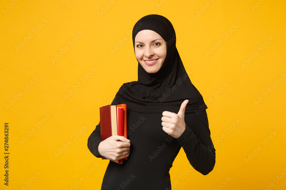 Arabian muslim student girl in hijab black clothes holds books isolated on yellow wall background, studio portrait. People religious lifestyle, education in high school concept. Mock up copy space. Arabian muslim student girl in hijab black clothes holds books isolated on yellow wall background, studio portrait. People religious lifestyle, education in high school concept. Mock up copy space.