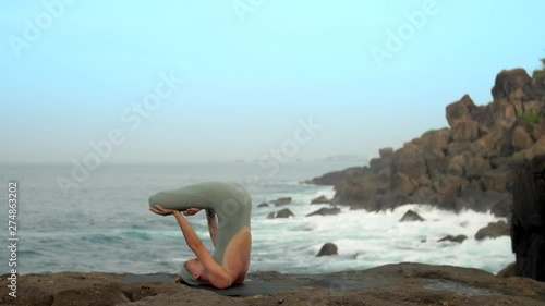 pretty slim woman stands in padma halasana on flat stone at boundless ocean coast under blue sky extreme slow motion. Concept yoga meditation and spiritual practices photo