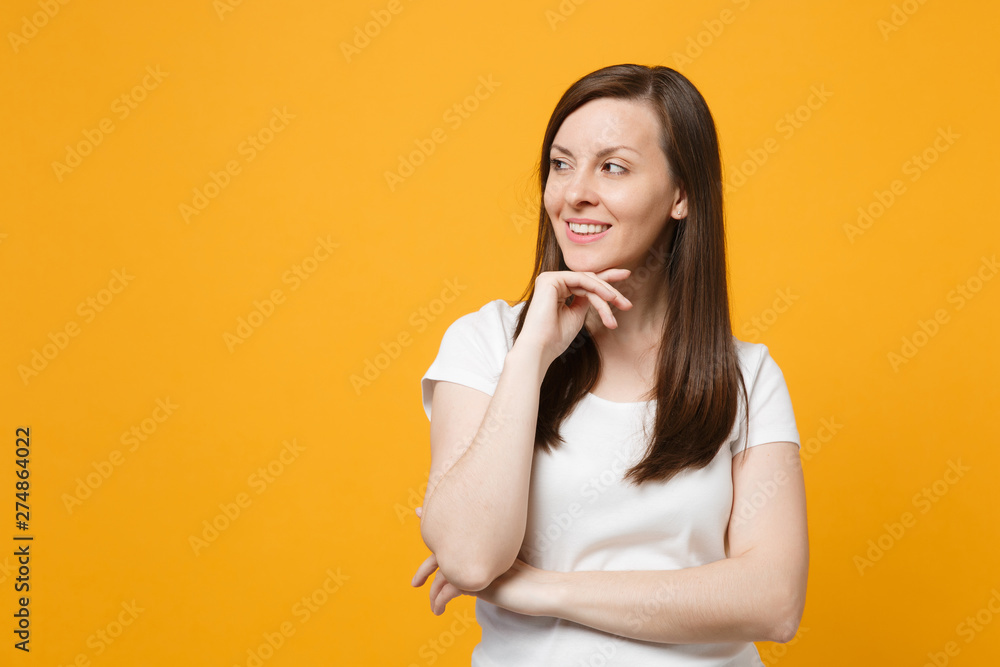 Portrait of smiling young woman in white casual clothes looking aside put hand prop up on chin isolated on bright yellow orange wall background in studio. People lifestyle concept. Mock up copy space. Portrait of smiling young woman in white casual clothes looking aside put hand prop up on chin isolated on bright yellow orange wall background in studio. People lifestyle concept. Mock up copy space.