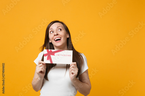 Portrait of pensive young woman in white casual clothes looking up, holding gift certificate isolated on bright yellow orange wall background in studio. People lifestyle concept. Mock up copy space.