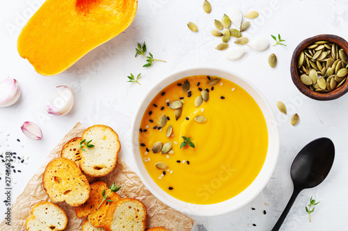 Autumn pumpkin cream soup in bowl served with seeds and crouton on white stone table top view.