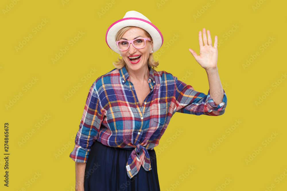 Portrait of surprised modern stylish mature woman in casual style with hat and eyeglasses standing waving her hand and looking at camera with toothy smile. studio shot isolated on yellow background. Portrait of surprised modern stylish mature woman in casual style with hat and eyeglasses standing waving her hand and looking at camera with toothy smile. studio shot isolated on yellow background.