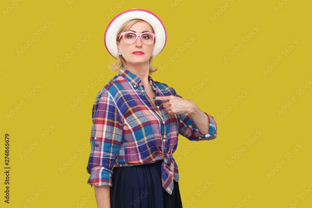 Because I am. Portrait of proud serious modern stylish mature woman in casual style with hat and eyeglasses standing, pointing herself and looking at camera. studio shot isolated on yellow background. Because I am. Portrait of proud serious modern stylish mature woman in casual style with hat and eyeglasses standing, pointing herself and looking at camera. studio shot isolated on yellow background.
