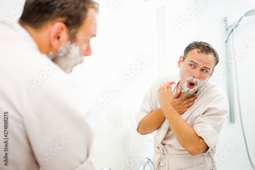 A young man in a bathrobe is shaving in the bathroom  looking in the mirror