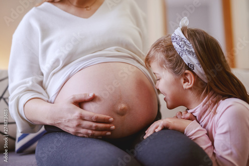 Cute little girl kissing mother's belly. Living room interior.