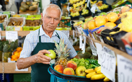 Senior male seller in apron putting fresh products on shelves in greengrocery