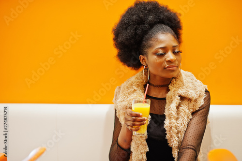 African woman sitting on cafe against orange wall with pineapple juice in hands.