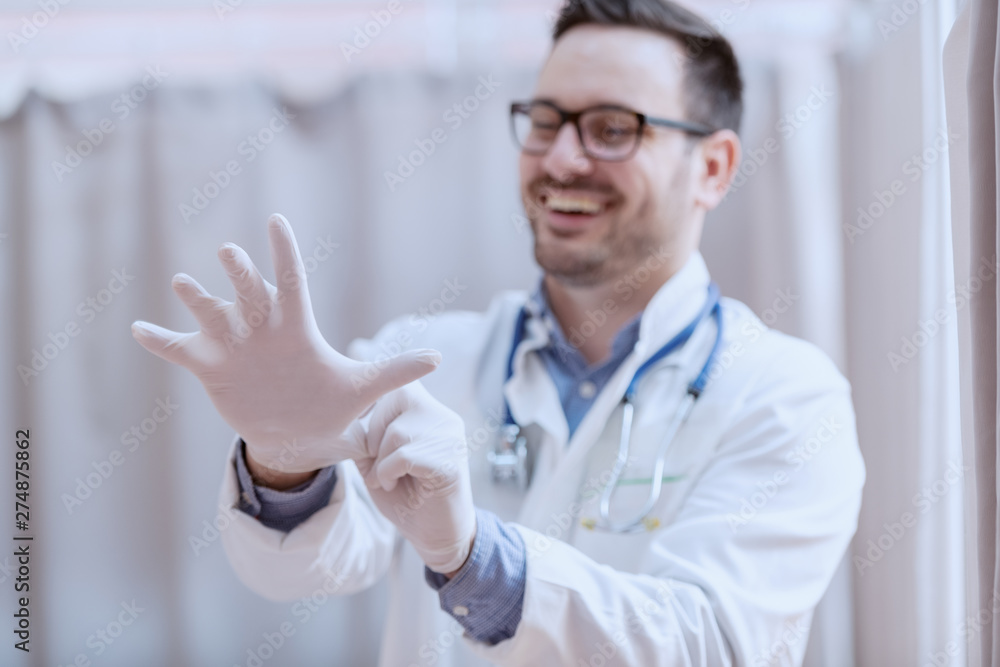 Young smiling Caucasian male doctor in white uniform, with eyeglasses and stethoscope around neck putting rubber gloves before patient examination. Selective focus on hands. Young smiling Caucasian male doctor in white uniform, with eyeglasses and stethoscope around neck putting rubber gloves before patient examination. Selective focus on hands.