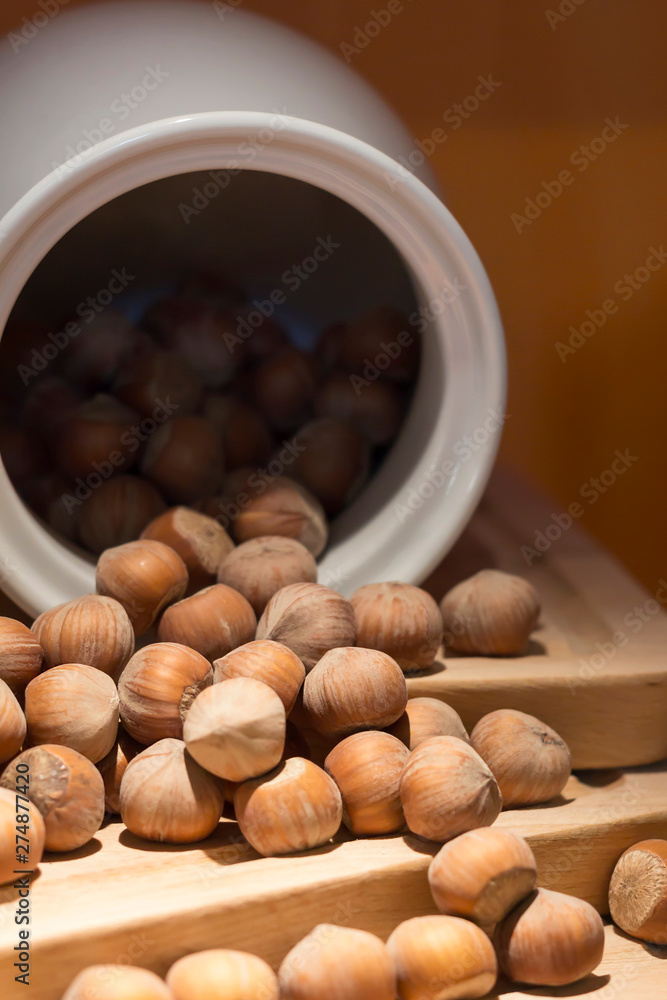 Hazelnut in shell poured from a white jug on wooden stand, brown orange background. Close-up, copy space, vertical layout Hazelnut in shell poured from a white jug on wooden stand, brown orange background. Close-up, copy space, vertical layout