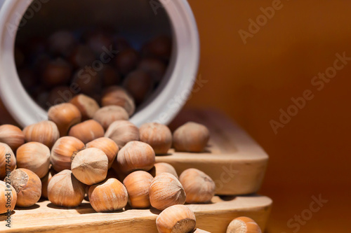 Hazelnut in shell poured from a white jug on wooden stand, brown orange background. Close-up, copy space
