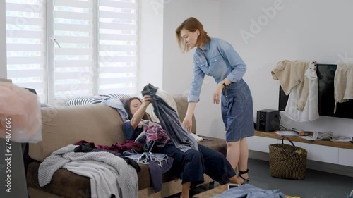 tired client girl is lying on couch in heap of clothes and her image-maker is standing near, taking gowns photo