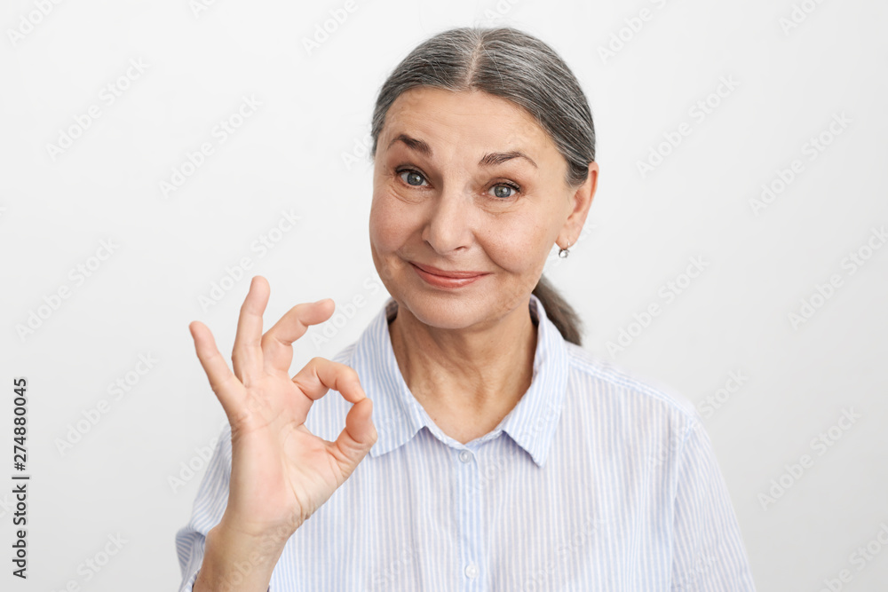 Picture of mature gray haired woman wearing blue shirt looking at camera with broad smile, showing okay gesture as sign of appreciation while helping daughter in law how to cook her speciality Picture of mature gray haired woman wearing blue shirt looking at camera with broad smile, showing okay gesture as sign of appreciation while helping daughter in law how to cook her speciality