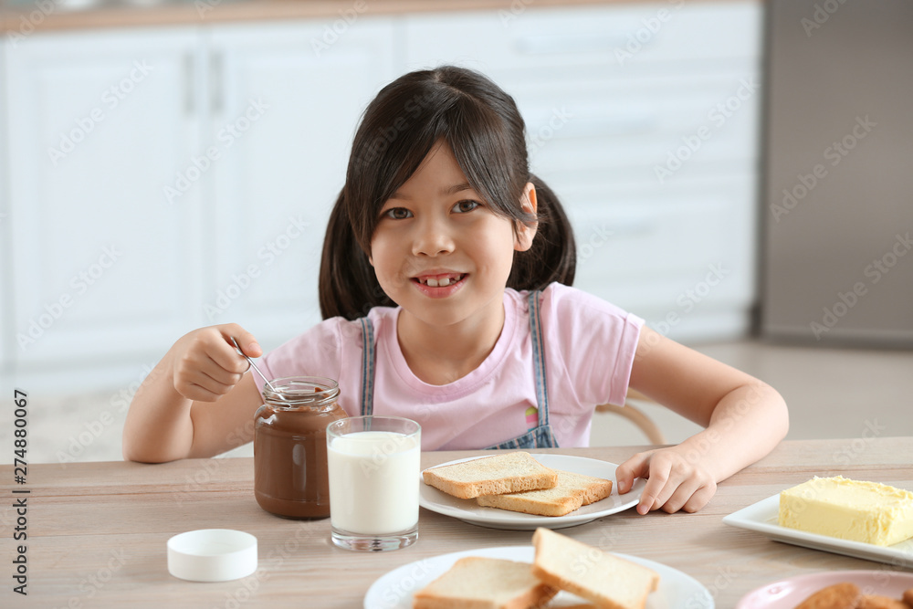 Funny little girl eating tasty toasts with chocolate spreading in kitchen