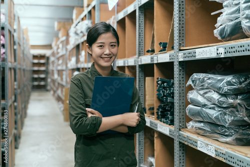 cute female worker holding clipboard in warehouse. young attractive girl staff standing in storehouse face camera smiling. happy woman stockroom employee surrounding by goods inventory in factory.