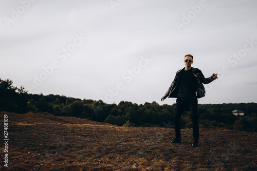 Young sexy man in leather jacket and sunglasses standing outdoor. Dark silhouette against grey sky