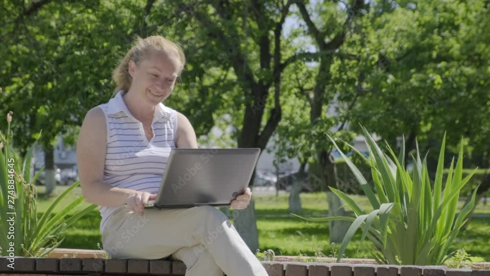 Happy woman communicates in video chat on laptop while sitting on park bench. girl laughs, smiles, shows sign like by hand looking at webcam. Happy woman communicates in video chat on laptop while sitting on park bench. girl laughs, smiles, shows sign like by hand looking at webcam.