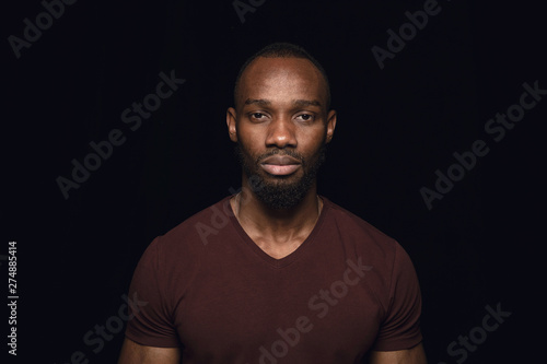 Close up portrait of young african man isolated on black studio background. Photoshot of real emotions of male model. Standing and looks serious. Facial expression, human nature and emotions concept.