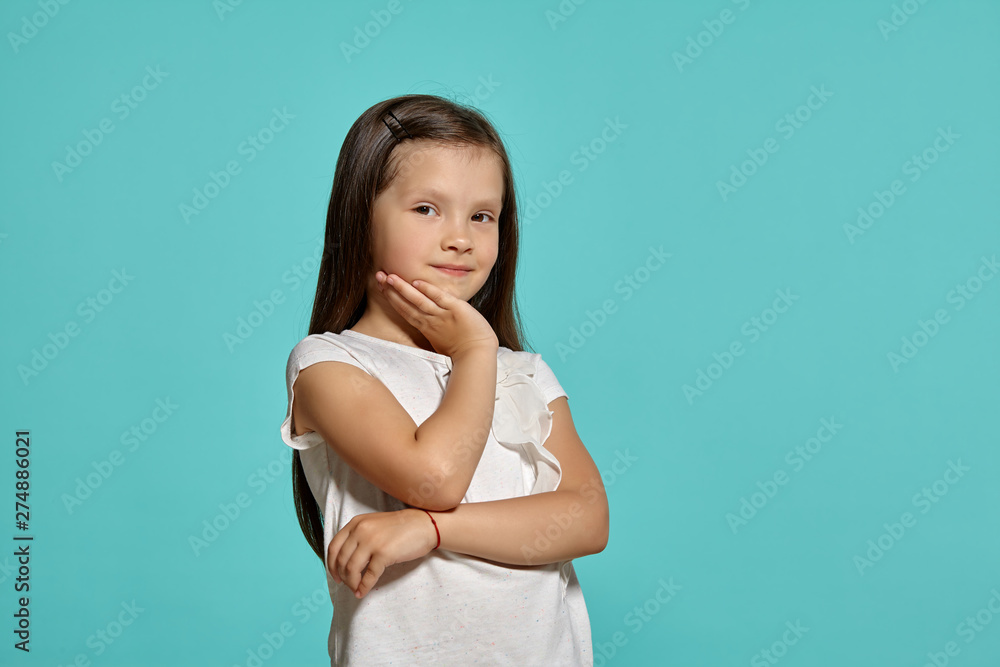 Close-up shot of a cute brunette little girl in a white blouse posing against a blue background. Close-up shot of a cute brunette little girl in a white blouse posing against a blue background.