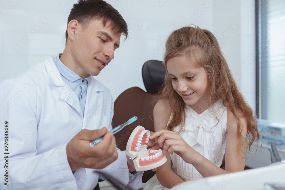 Charming young girl learning brushing teeth at dental clinic. Professional dentist teaching little girl brushing teeth, using dental model and a toothbrush Charming young girl learning brushing teeth at dental clinic. Professional dentist teaching little girl brushing teeth, using dental model and a toothbrush