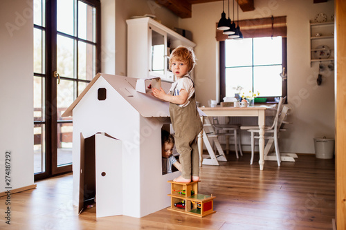 Two toddler children playing with a carton paper house indoors at home.