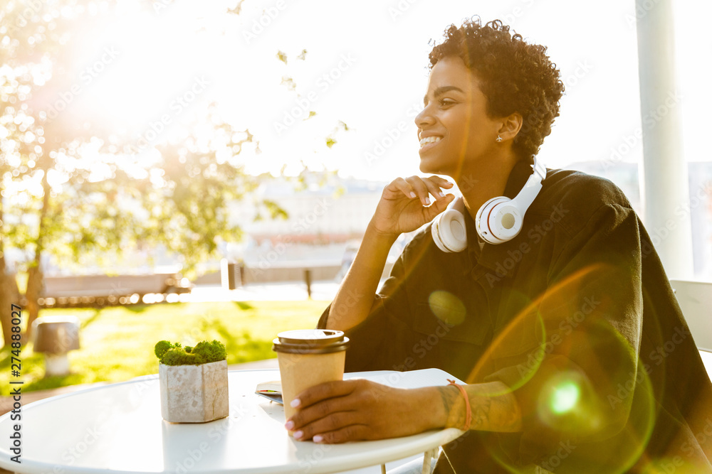 Fototapeta premium Portrait of cute african american woman with headphones holding paper cup while sitting in street cafe