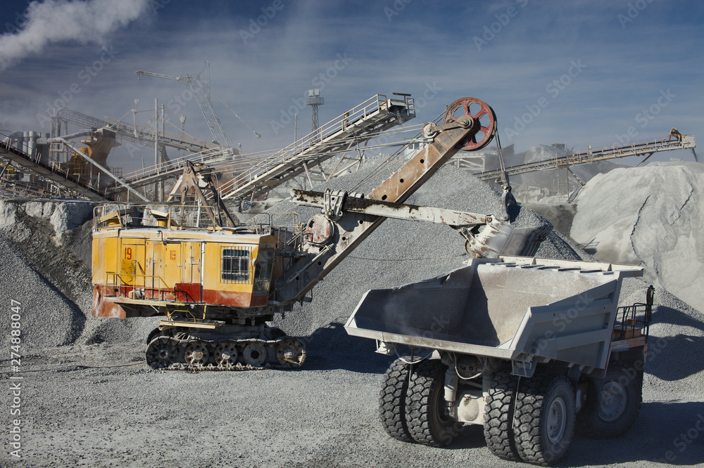 Heavy truck drives up to the excavator for gravel loading on the background of a rock stone crushing complex. Mining industry. Quarry mining equipment. Heavy truck drives up to the excavator for gravel loading on the background of a rock stone crushing complex. Mining industry. Quarry mining equipment.