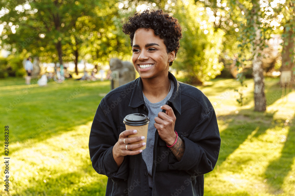 Photo of young african american woman holding and drinking takeaway coffee from paper cup while walking in city park Photo of young african american woman holding and drinking takeaway coffee from paper cup while walking in city park