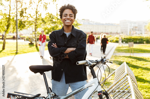 Photo of young african american woman standing with bicycle while walking in city park