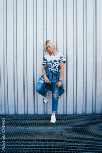 Beautiful girl with short white hair dressed in jeans in urban style stands on the background of a white wall. Her hair cover half face. She is looking forward A look at the camera © Andriy