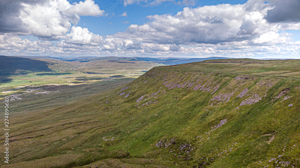 An aerial view of a mountain valley with green slope, railway bride and mountain summit range in the background under a majestic blue sky and huge altitude white clouds An aerial view of a mountain valley with green slope, railway bride and mountain summit range in the background under a majestic blue sky and huge altitude white clouds