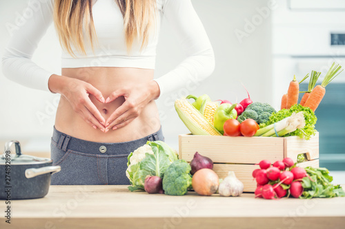 Woman with sports figure on her belly shows heart shape in home kitchen with wooden box full of organic vegetable