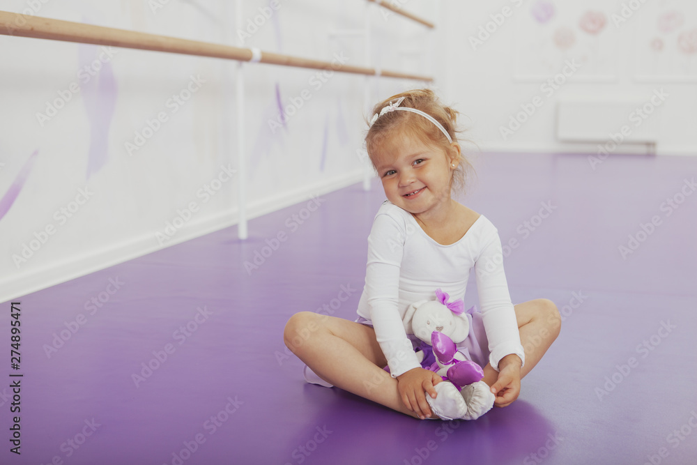 Adorable little ballerina smiling to the camera happily, sitting on the floor at dance studio with her toy. Cute little girl resting after dancing class