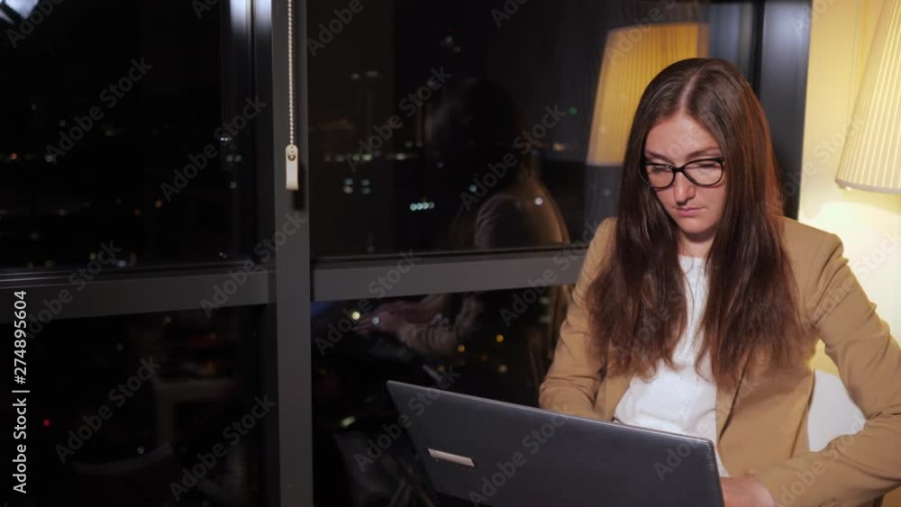 Overtime job of young woman in modern office with panoramic city view at night. She is sitting and working on her computer.
