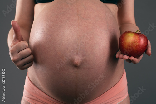 Pregnant woman is holding in hand a red apple and a thumbs up on a gray background. photo