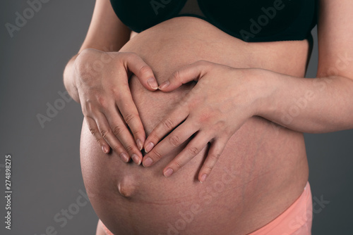 Pregnant woman is showing a heart shape by her fingers on a her belly background.