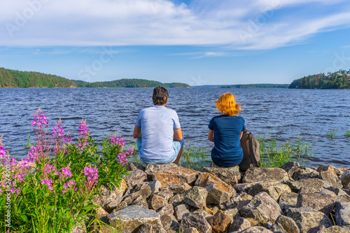 Tourists dark-haired man and redhead young woman sitting on stones on shore of lake bay in sunny summer evening. People resting and admiring beautiful landscape of Ladoga lake. Karelia, Russia