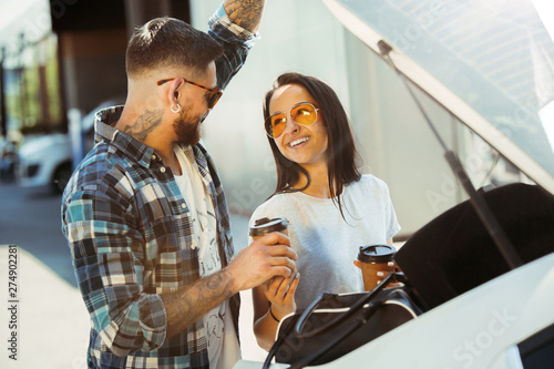 Young couple preparing for vacation trip on the car in sunny day. Woman and man drinking coffee and ready for going to sea or ocean. Concept of relationship, vacation, summer, holiday, weekend.