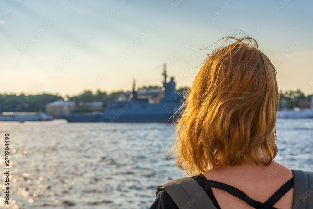 Redhead tween tourist girl on Neva river embankment looking at warships and architectural ensemble in summer evening at sunset. Authentic lifestyle moments, travel concept. Saint Petersburg, Russia