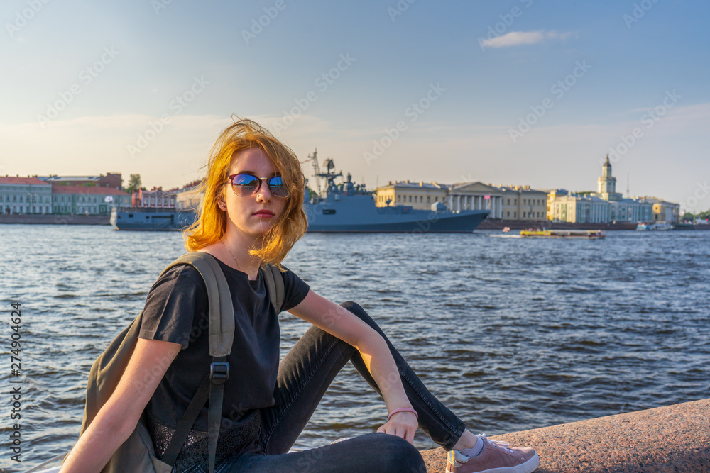 Redhead teen tourist girl on Neva river embankment posing against warships and architectural ensemble in summer evening at sunset. Authentic lifestyle moments, travel concept. Saint Petersburg, Russia