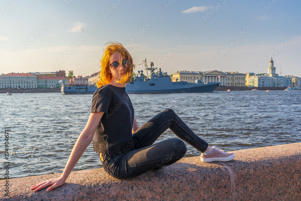 Redhead teen tourist girl on Neva river embankment posing against warships and architectural ensemble in summer evening at sunset. Authentic lifestyle moments, travel concept. Saint Petersburg, Russia